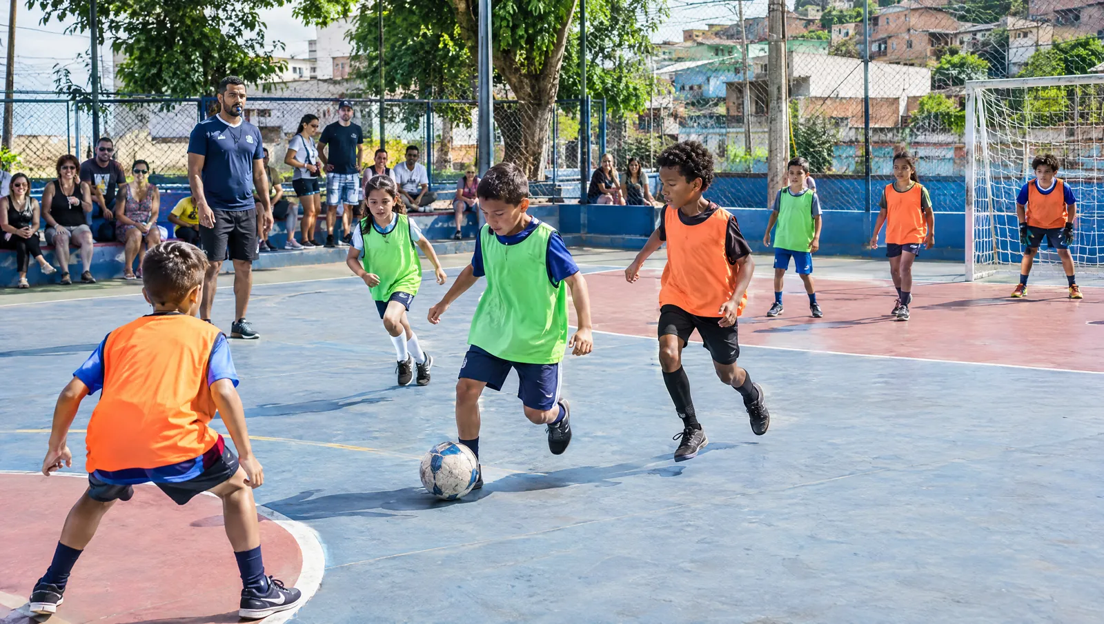 Numa quadra de escola num sábado de sol, crianças de colete jogam futebol enquanto os pais assistem da lateral